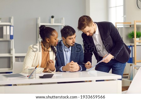 Bank employee gives documents for signature to a Caucasian man and his dark-skinned wife. Specialist in lending and financial assistance for young families indicates the place of signature.