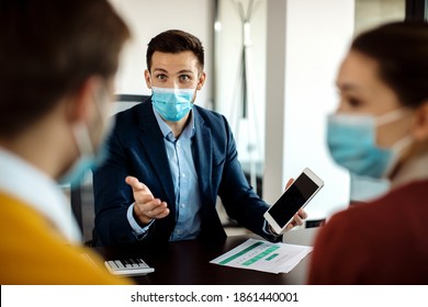 Bank Manager Wearing Face Mask And Communicating With A Couple While Using Digital Tablet On A Meeting In The Office. 