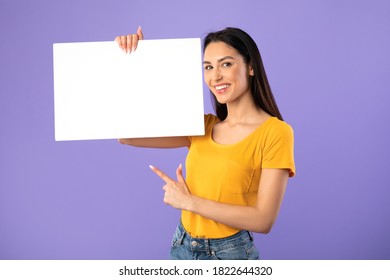 Banner For Advertising. Portrait Of Smiling Lady Holding White Empty Square Placard In Hand And Pointing At It, Isolated On Purple Studio Background, Empty Space. Young Woman Showing Poster For Text