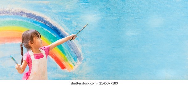 Banner of asian little girl is painting the colorful rainbow and sky on the wall and she look happy and funny, concept of art education and learn through play activity for kid development.