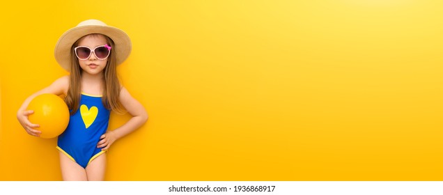 Banner With Caucasian Happy Little Kid In A Blue Swimsuit On A Yellow Background. A Cute Girl In A Model Pose Is Smiling In A Blue Bathing Suit, Hat And Sun Glasses, Holding A Volleyball. Copyspace