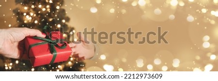 Banner with Christmas gift on beige bokeh background. Xmas greeting card with copy space. Hands of parent giving gift to child.