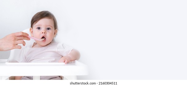 Banner copyspace baby girl eating blend mashed food sitting, on high chair, mother feeding child, hand with spoon for vegetable lunch, baby weaning, first solid food for young kid. - Shutterstock ID 2160304511