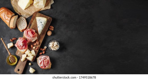 Banner with delicious snack of prosciutto, cheese, nuts and honey. Wooden chopping board. Dark textured background. Appetizer, aperitif, snack table. Top view. Space for text. - Powered by Shutterstock