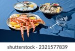 Banner with a gourmet seafood platter of prepared crab phalanges, lemon slices, sauces, and fresh oysters on ice. Set on a blue tablecloth with a solid blue backdrop casting stark shadows.