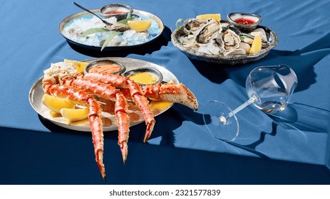 Banner with a gourmet seafood platter of prepared crab phalanges, lemon slices, sauces, and fresh oysters on ice. Set on a blue tablecloth with a solid blue backdrop casting stark shadows. - Powered by Shutterstock