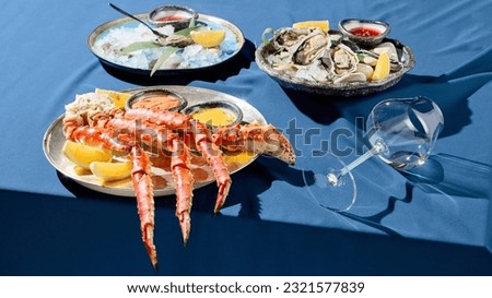 Banner with a gourmet seafood platter of prepared crab phalanges, lemon slices, sauces, and fresh oysters on ice. Set on a blue tablecloth with a solid blue backdrop casting stark shadows.