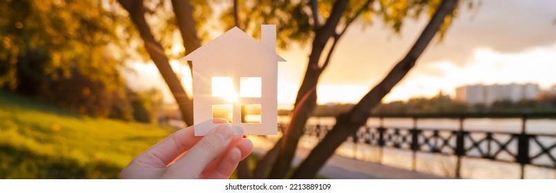 Banner With Hand That Holds A Paper House Against The Background Of The Sunset Sky. The Sun Is Setting Over The Horizon. The Concept Of Your Own Home, Personal Space And Mortgage. High Quality Photo