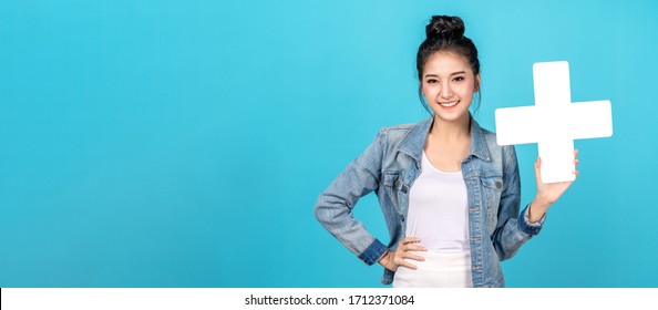 Banner of Happy asian woman smiling and holding plus or add sign on copy space blue background. Cute asia girl smiling wearing casual jeans shirt and showing join sign for increse and more concept