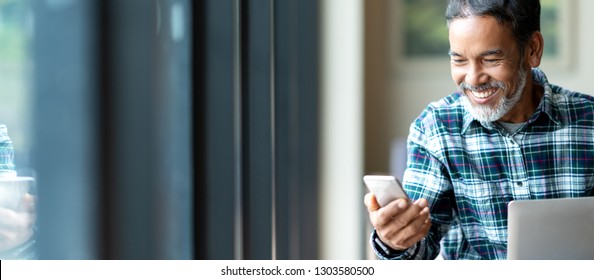 Banner Of Happy Mature Asian Or Hispanic Old Man Looking At Smartphone Smiling Positive In Casual Lifestyle With Digital Technology Of Older Or Senior Guy Concept. Urban Elderly Asian Retired Man.