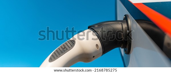Banner image. power cord for electric car. Green\
station.Power supply for electric car battery charging.Blue\
sky.Selective focus.Closeup.Copy\
space.