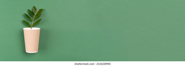 Banner with kraft paper coffee cup with green leaves - biodegradable, compostable paper utensils for hot drinks. Paper cup on green background with copy space. Environmental conservation concept
