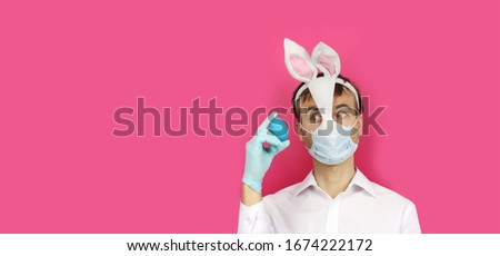 banner with man in mask Easter Bunny and medical mask look to blue egg, virus or coronavirus protection and prevention before holiday, preparation for party, copy space with pink background   