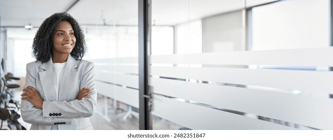 Banner portrait of 30s latin business woman with crossed arms looking aside. Confident smiling young African American middle age leader female businesswoman on modern office background, copy space. - Powered by Shutterstock