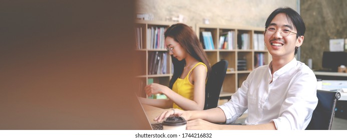 Banner Portrait Of Young Happy Millenials Asian Male Worker Sitting On Desk Table Smile To Camera At Office In Concept Future Of Work Stay Six Feet Away, Post Covid New Normal Workplace Or Workspace.