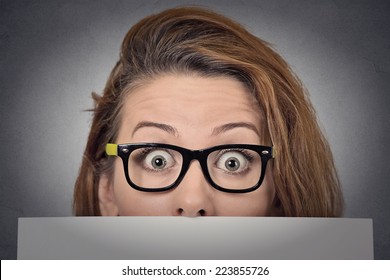 Banner sign woman peeking over edge of blank empty paper billboard. Beautiful young woman with glasses looking surprised and scared funny with wide open eyes isolated on grey wall background