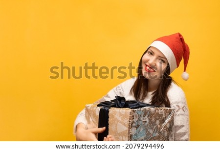 Banner with smiling brunette in red Santa Claus hat and warm white sweater with big Christmas gift. Happy girl isolated against yellow background holds New Year silver present box with blue ribbon.