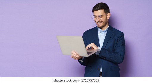 Banner of smiling handsome businessman wearing eyeglasses, using touchpad of open laptop he is holding in hands, isolated on purple background with copyspace on left