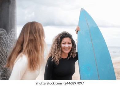 A banner with two young female surfers talking next to their surfboards. Portrait of a Latino woman with curly hair who smiles, her face has colorful zinc and she holds a blue surfboard with her hand. - Powered by Shutterstock