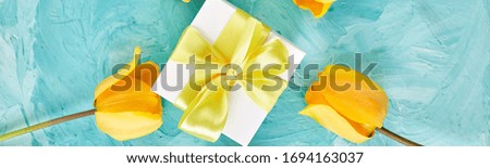 Banner of White gift box with yellow ribbon near yellow tulip on blue background. Flat lay. Mother or Woman Day. Greeting Card. Copy space. Spring.