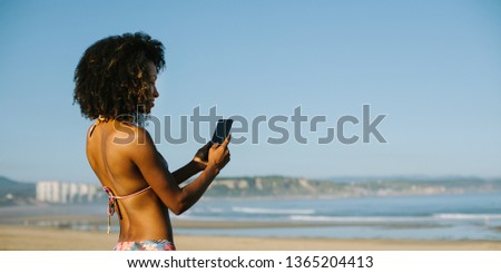 Banner of young beautiful black woman texting on cell phone at the beach on summer or spring vacation. Mobile communication on travel concept. San Juan de Nieva, Asturias, Spain.