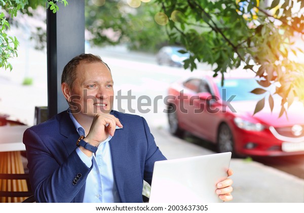 Banner with a young male businessman with a smile waits\
for a business partner, a Caucasian in a blue suit sitting at a\
table in a summer cafe with a laptop. Background street and red car\
in blur. 