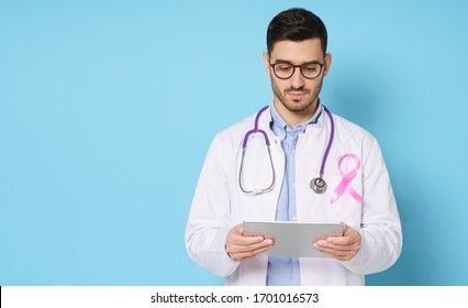 Banner Of Young Oncologist Doctor With Pink Ribbon On His White Coat Looking At Tablet, Using App For Medical Check Up, Isolated On Studio Blue Background. Breast Cancer Awareness Month Concept