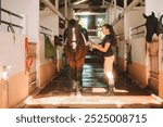 Banner with young woman rider in equestrian uniform in stable saddling her horse. Horse riding training. She throws saddle on brown horse