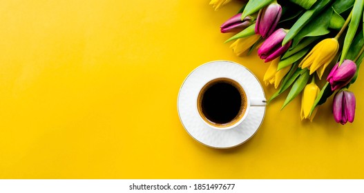 Banner.A cup of hot, morning coffee and a bouquet of yellow-lilac tulips on a bright yellow background. View from above. Close-up. Copy space for text. The concept of holidays and good morning wishes.