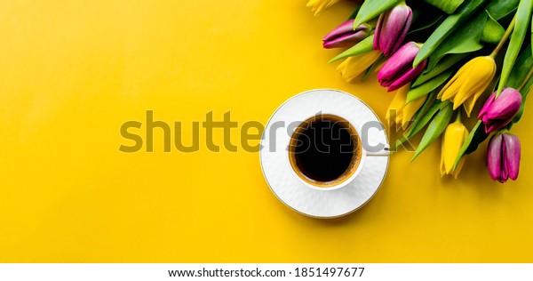 Banner.A cup of hot, morning coffee and a bouquet\
of yellow-lilac tulips on a bright yellow background. View from\
above. Close-up. Copy space for text. The concept of holidays and\
good morning wishes.