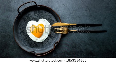 Banner.Homemade fried egg in a vintage pan and stylish cutlery on a dark background. Heart-shaped yolk.View from above. Copy space for text.