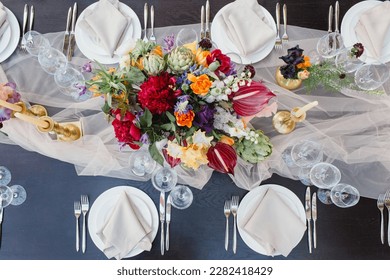 Banquet table setting in blue and white colors, top view. In the center of the table is a bouquet of bright exotic flowers on a drapery. Candles in gold candlesticks, plates with napkins, glasses.