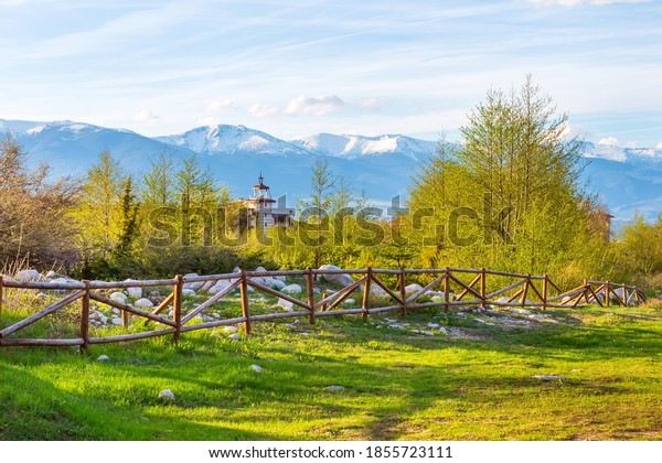 Bansko, Bulgaria\
spring landscape with the wooden fence, trees, tower of chalet and\
snowy Rila mountains\
peaks