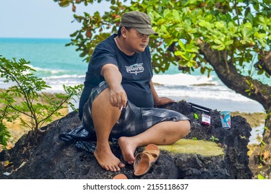 BANTEN, INDONESIA - MAY 7, 2022: An Asian Adult Man In A Gray Hat Sitting Relaxing On A Rock By The Beach Looking Ahead. Family Picnic On The Beach At Pulo Manuk, Banten