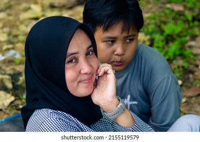 BANTEN, INDONESIA - MAY 7, 2022: Close Up Of A Young Asian Muslim Woman Looking At The Camera Wearing A Black Headscarf Sitting Cheerfully With A Small Child. Family Picnic On Pulo Manuk Beach, Banten