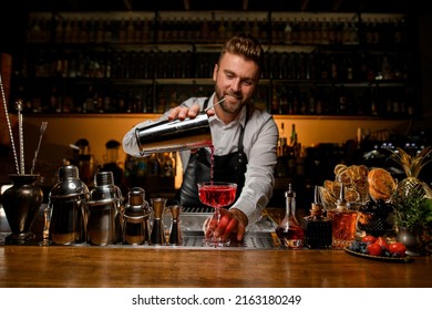 bar counter with a variety of bar equipment and a transparent crystal goblet in which the bartender pours a cocktail from a steel shaker cup
