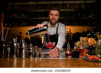 bar counter with a variety of bar equipment and a transparent crystal goblet in which the bartender pours a cocktail from a steel shaker cup