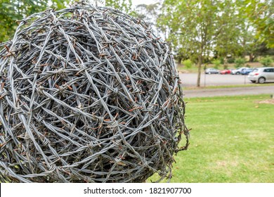 Barb Wire Rolled Up Into A Ball Shaped Ornament, Anchored Onto A Fence In A Public Park.