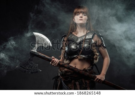 Barbaric beutiful woman from north armed with two handed axe and with brown hairs poses in dark foggy background.