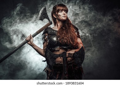 Barbaric female viking in light armour with brown hairs poses in dark smokey background holding two axes.