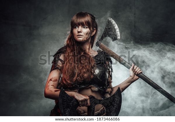 Barbaric female viking in
light armour with brown hairs poses in dark smokey background
holding two axes.