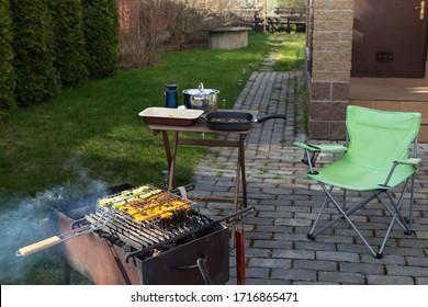 Barbecue With Grilled Vegetables With Smoke. Folding Chair And A Small Table. House Porch And Backyard In The Background. Spring. Summer