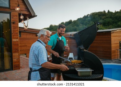 Barbecue. Leisure, food, family and holidays concept. Senior father and son wearing aprons, drinking beer and roasting meat on barbecue in the backyard