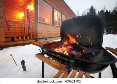 Barbecue stove with burning coals at winter evening near wooden cottage