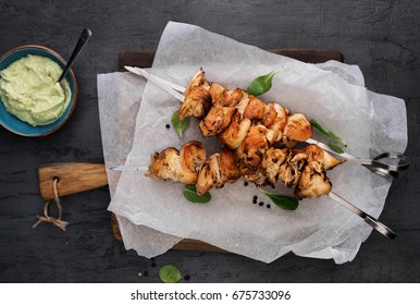 Barbecued chicken breast skewers with avocado sauce on dark background, top view