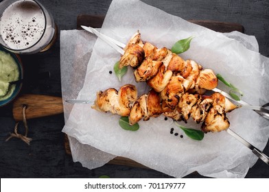 Barbecued chicken breast skewers with avocado sauce and beer on dark wooden table close up, top view