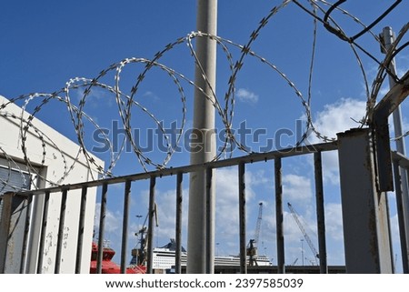 The barbed wire on top of the fence enclosing the passenger dock. The fence protects against unauthorized entry. Behind are visible superstructures of different vessels.