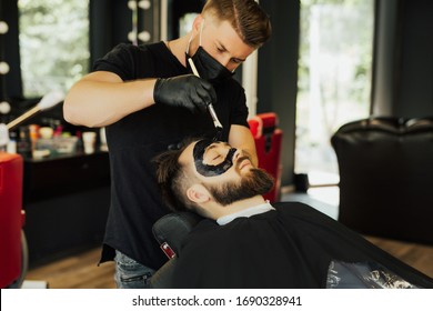 Barber Applies Black Charcoal Mask To Man Face To Clean Pore Skin And Remove Acne From Nose In Barber Shop. Spa And Skin Care For Men. Young Male Lying On Chair In Barber Shop.