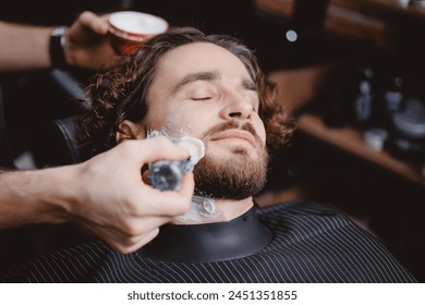 Barber applies shaving foam with brush to man beard in barbershop, warm toning. - Powered by Shutterstock