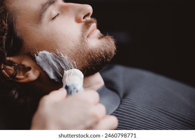 Barber applies shaving foam with brush to man beard in barbershop, warm toning, top view